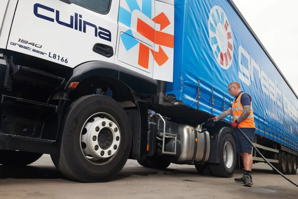 Front angled image of a man in orange hi-vis filling a white Cuilna lorry with a blue trailer, up with fuel
