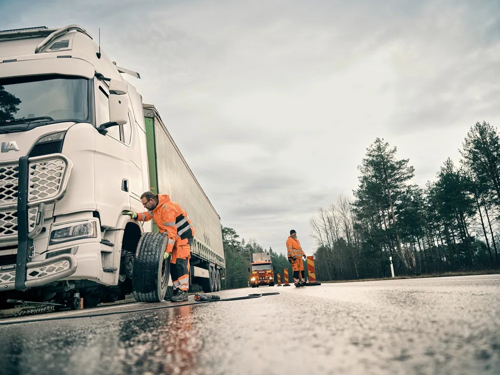 Man in an orange hig vis suit replacing a front wheel on a lorry. The road is wet and there is a red lorry in the background