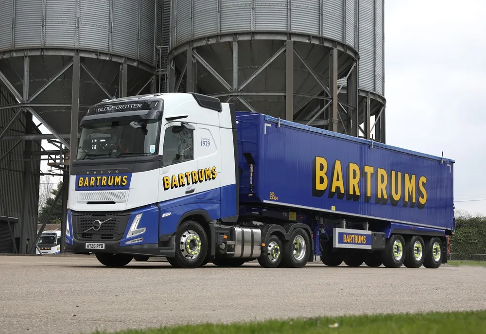 Image of a white truck with a blue trailer labelled Bartrums from a front angled and side view parked on concrete in front of silos.